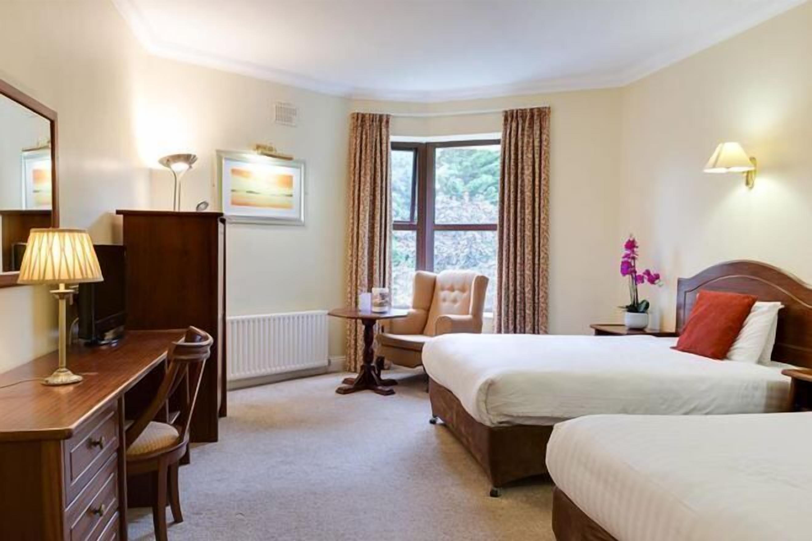 A hotel room with a bed, a desk, a chair, a lamp, a window with curtains, and a wall-mounted lamp.