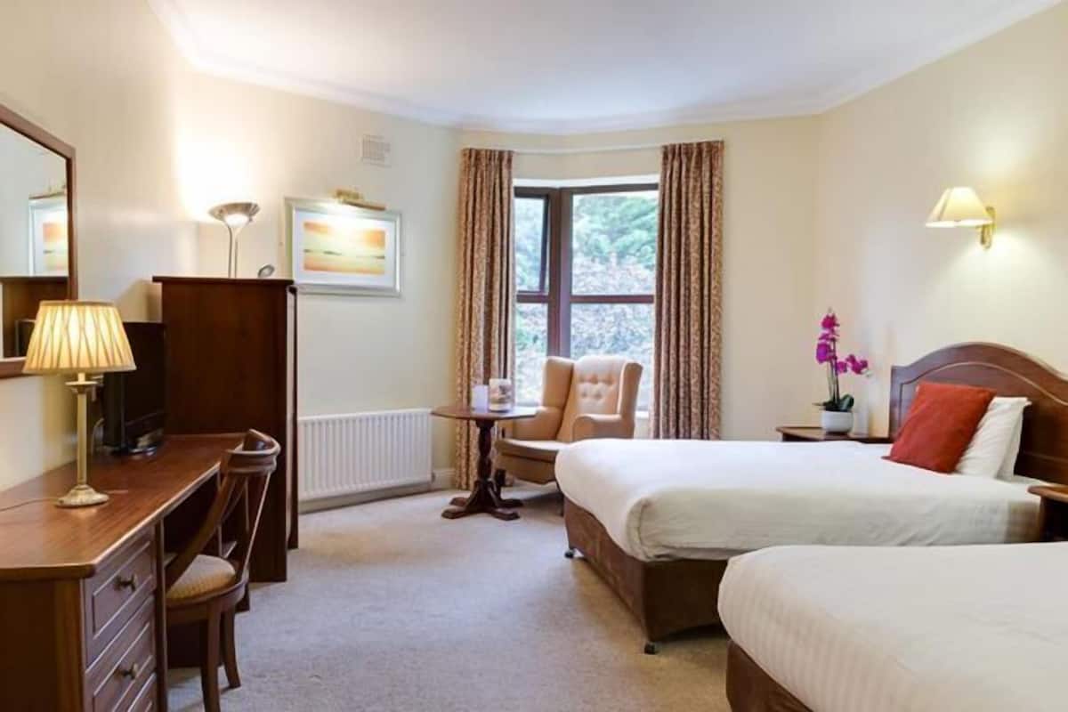 A hotel room with a bed, a desk, a chair, a lamp, a window with curtains, and a wall-mounted lamp.