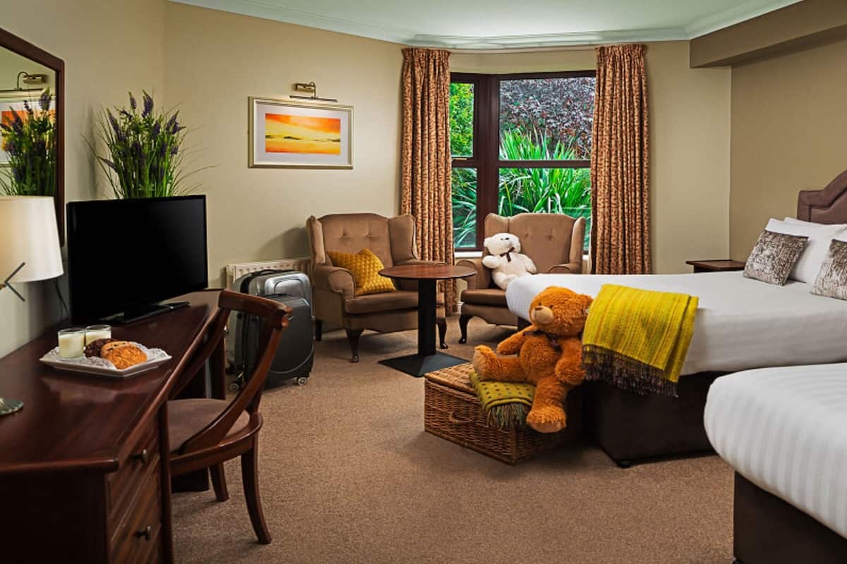 A hotel room with a bed, a chair, a desk with a television, and a teddy bear.