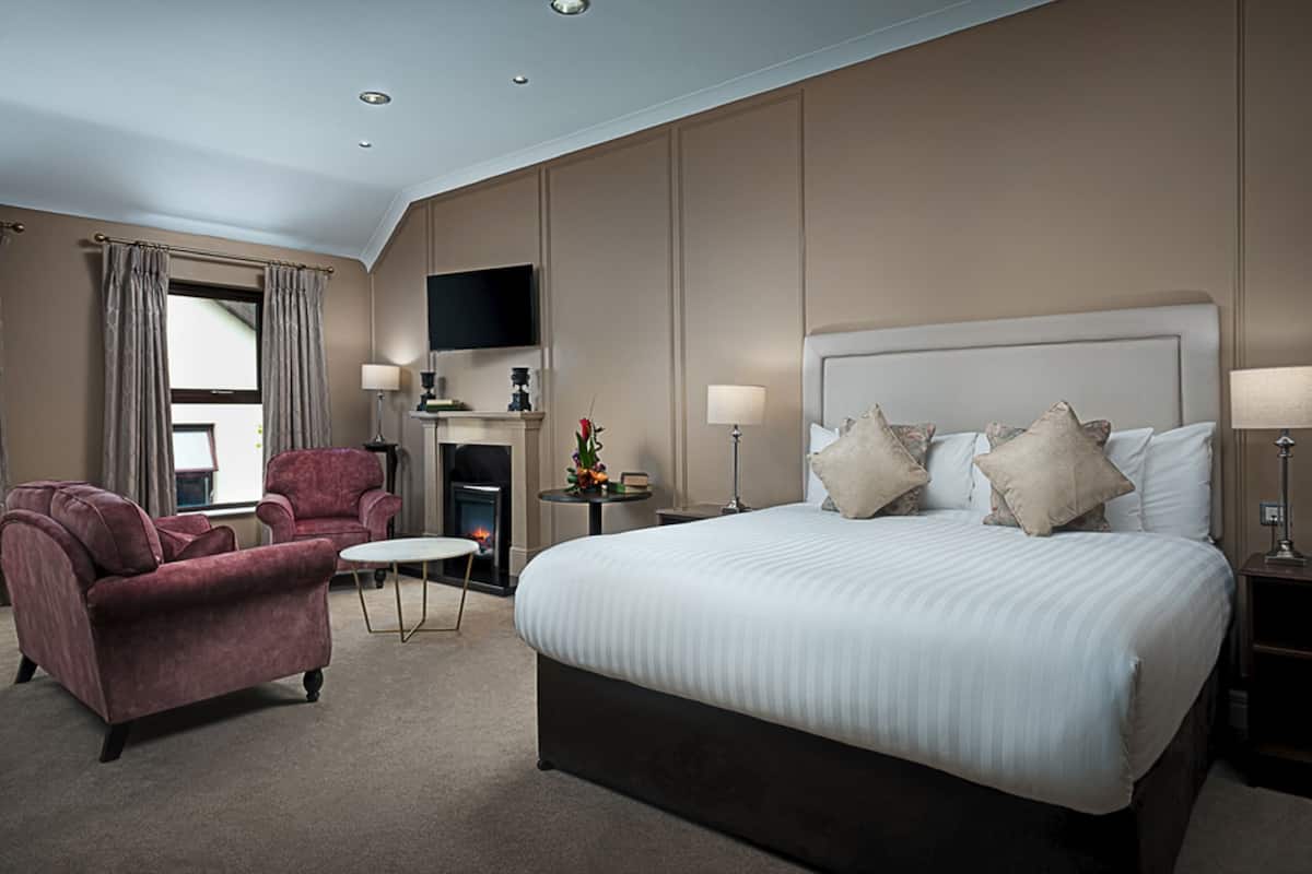 A hotel room with a large bed, two armchairs, a small table, a fireplace, and a TV mounted on the wall.