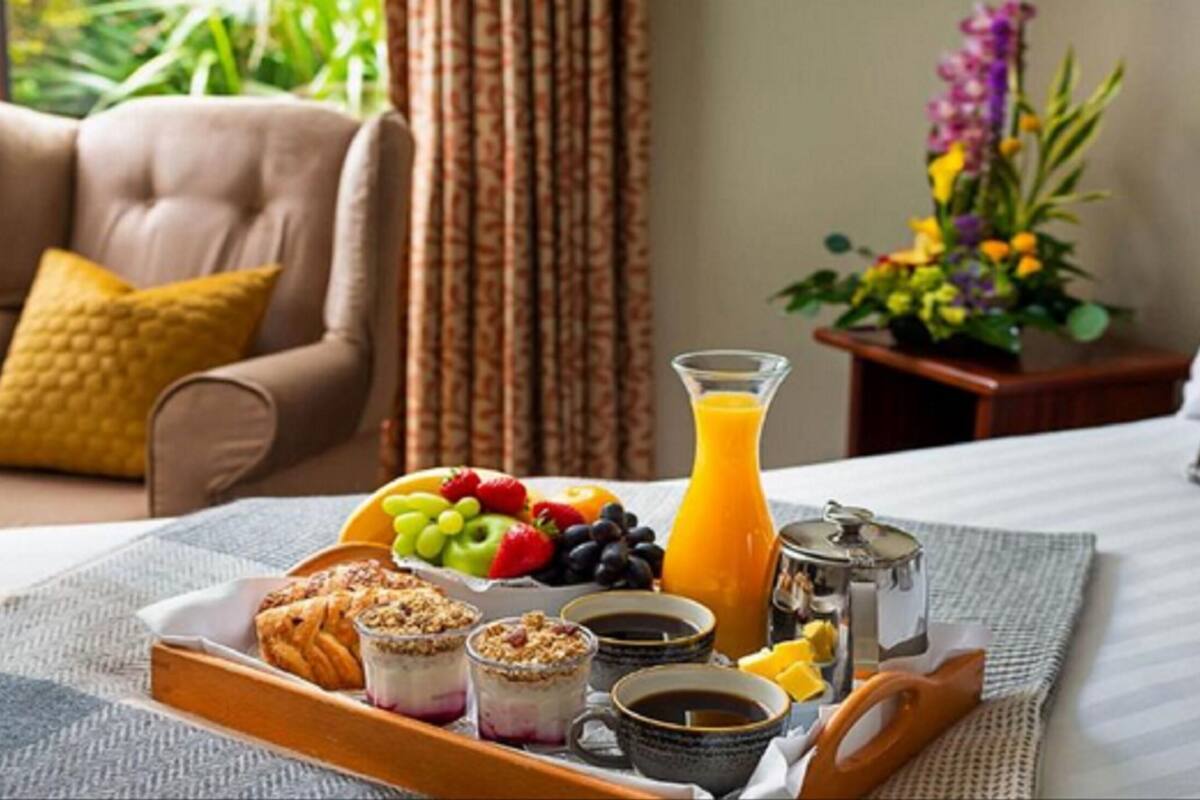 A breakfast tray with fruits, juice, pastries, and coffee on a bed with a view of greenery.