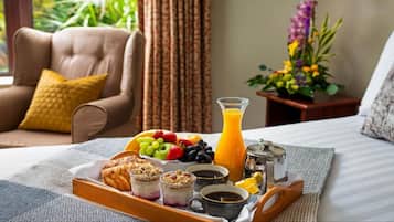 A breakfast tray with fruits, juice, pastries, and coffee on a bed with a view of greenery.
