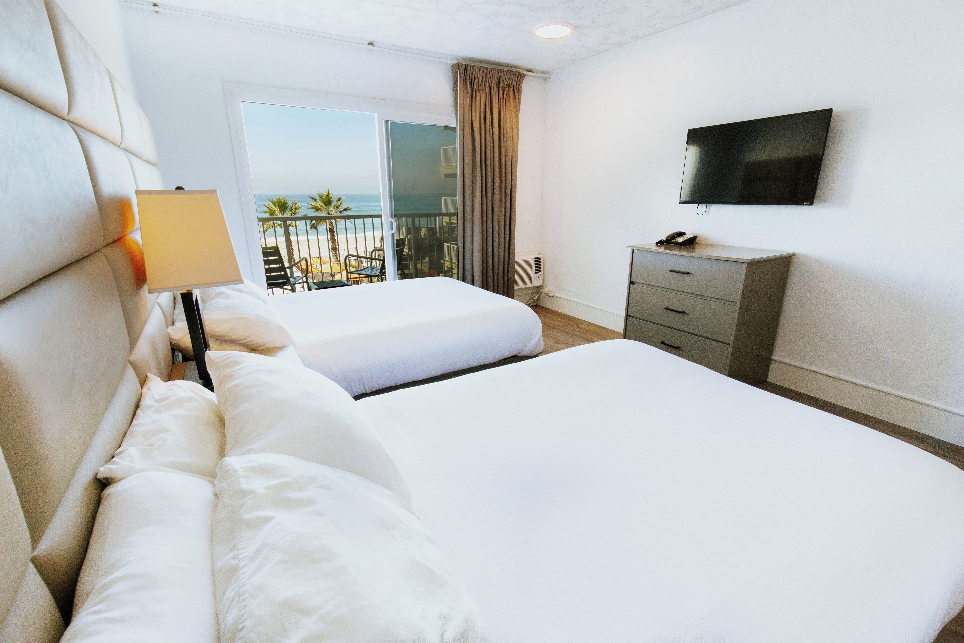 A hotel room with two beds, a TV, a desk, and a balcony with a view of the beach.