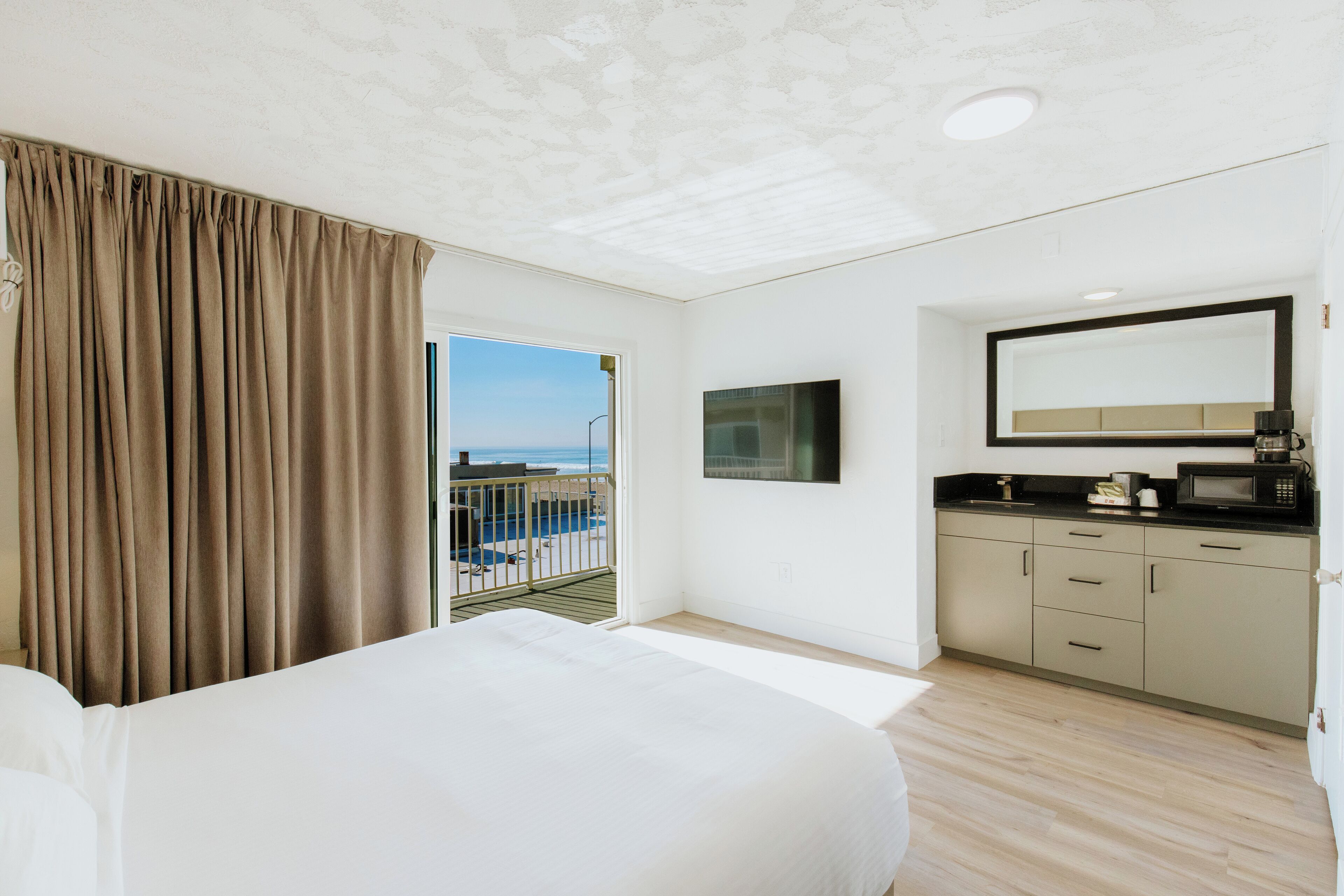 A hotel room with a large bed, a kitchenette, a balcony with a view of the sea, and a ceiling with a textured pattern.