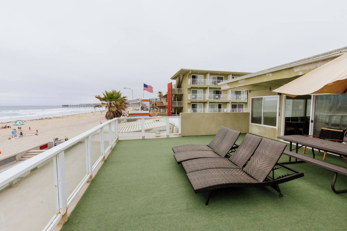 A balcony with a green artificial turf, a wicker sofa, a picnic table, and a view of the beach and ocean.
