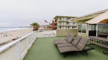A balcony with a green artificial turf, a wicker sofa, a picnic table, and a view of the beach and ocean.