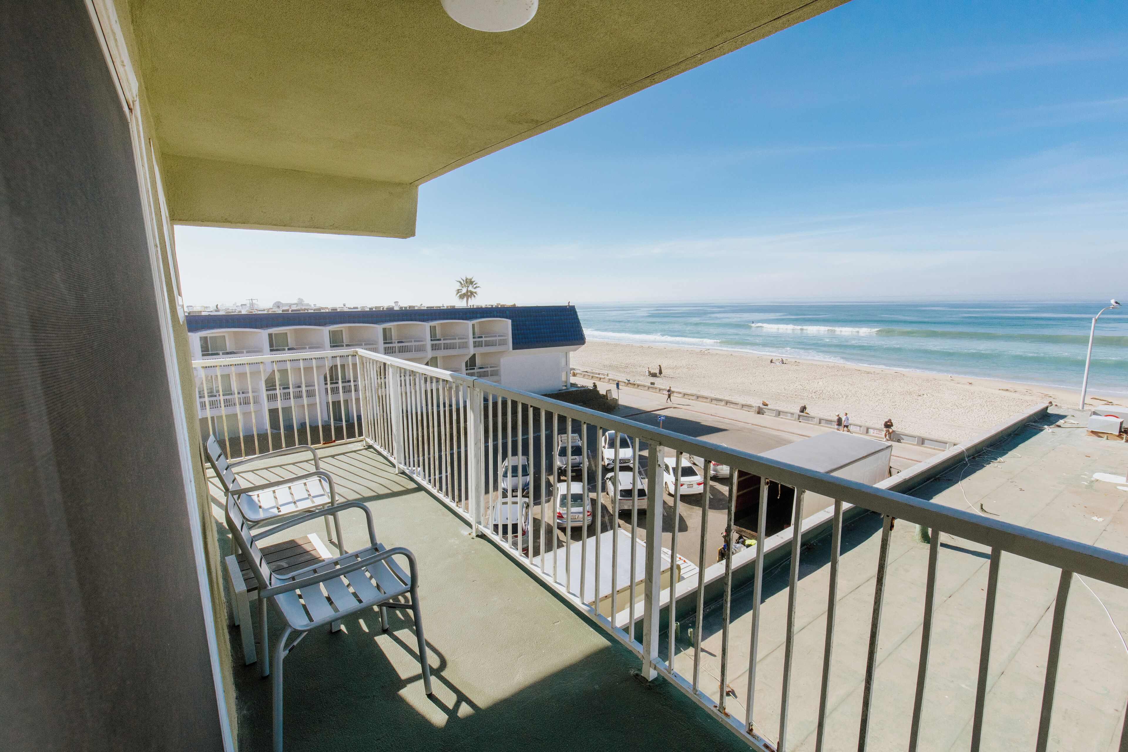 A balcony with a view of the beach, a table, chairs, and a parking area.