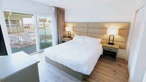 Hotellihuone, jossa on sänky, kaksi yöpöytää, yölamppu, liukuovi parvekkeelle, jossa on ulkotilaan sijoitettuja istuimia, sekä näkymä ulos.