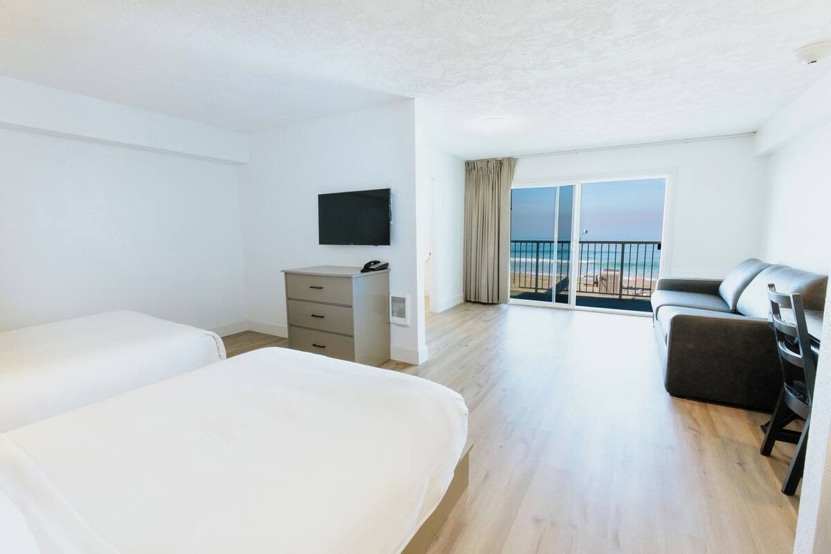 A hotel room with two beds, a TV, a desk, and a balcony with a view of the beach.
