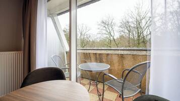 Een balkon met een tafel en twee stoelen, uitzicht op bomen en een gebouw op de achtergrond.