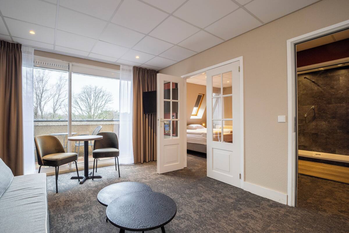 Ein Hotelzimmer mit einer Couch, zwei Sesseln, einem kleinen Tisch, einem Bett und einem Duschbereich.