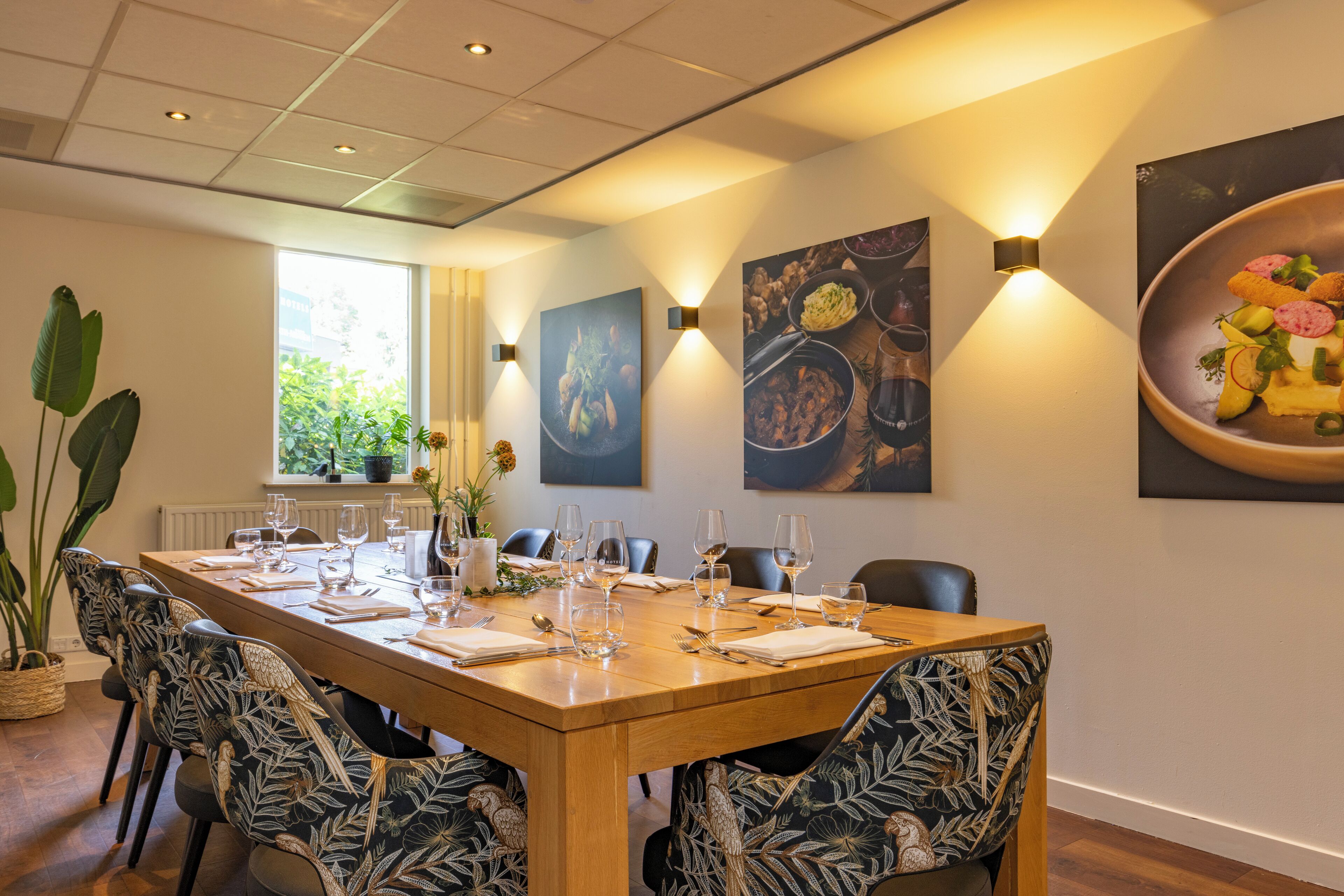 A dining area with a long wooden table set for six, surrounded by chairs with a leaf pattern upholstery. There are wall-mounted light fixtures and framed artwork above the table.