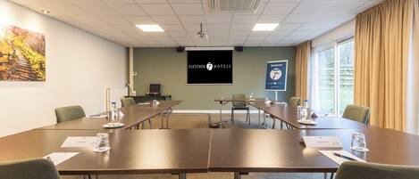 Une salle de conférence équipée d’une longue table, de chaises, d’un écran de projection et d’une banderole de bienvenue.