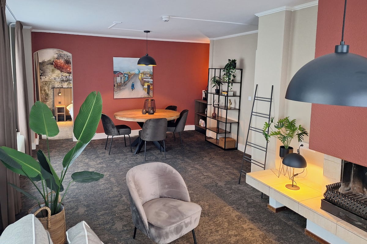 Ein modernes Wohnzimmer mit einem runden Holztisch, grauen Stühlen, einer schwarzen Pendelleuchte, einem Bücherregal und einem Kamin.