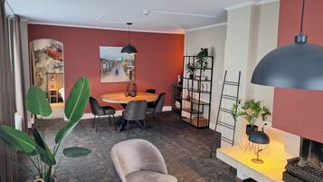 A modern living room with a round wooden table, grey chairs, a black pendant light, a bookshelf, and a fireplace.