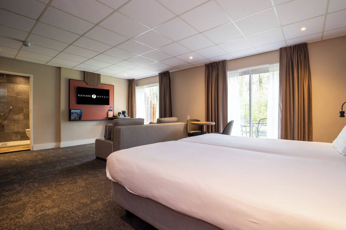 Ein Hotelzimmer mit einem großen Bett, einer Sitzecke mit Tisch, einem an der Wand befestigten Fernseher und Blick ins Freie durch die Fenster.