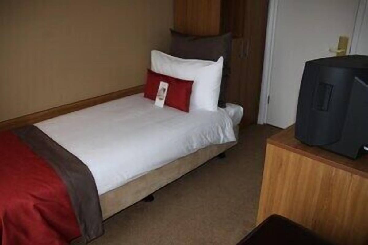 Ein Einzelbett mit weißer und roter Bettdecke, ein Fernseher auf einem Holztischchen und ein Holzschrank in einem Hotelzimmer.