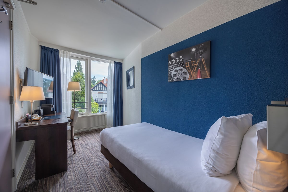 Habitación de hotel con cama, escritorio y silla, una ventana con cortinas y una pared de acento azul.