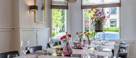 Un comedor con mesas dispuestas para dos, cristalería transparente y jarrones con flores.