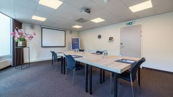 Een vergaderruimte met een lange tafel, stoelen, een projecter, een scherm, een klok, een telefoon en een welkomstbanner.