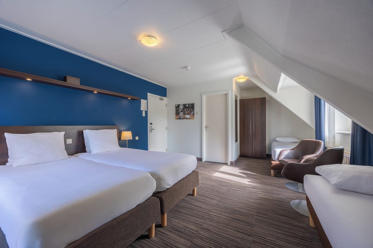 Habitación de hotel con dos camas, una zona de estar con una silla, un estante de madera y una puerta que da a otra habitación.