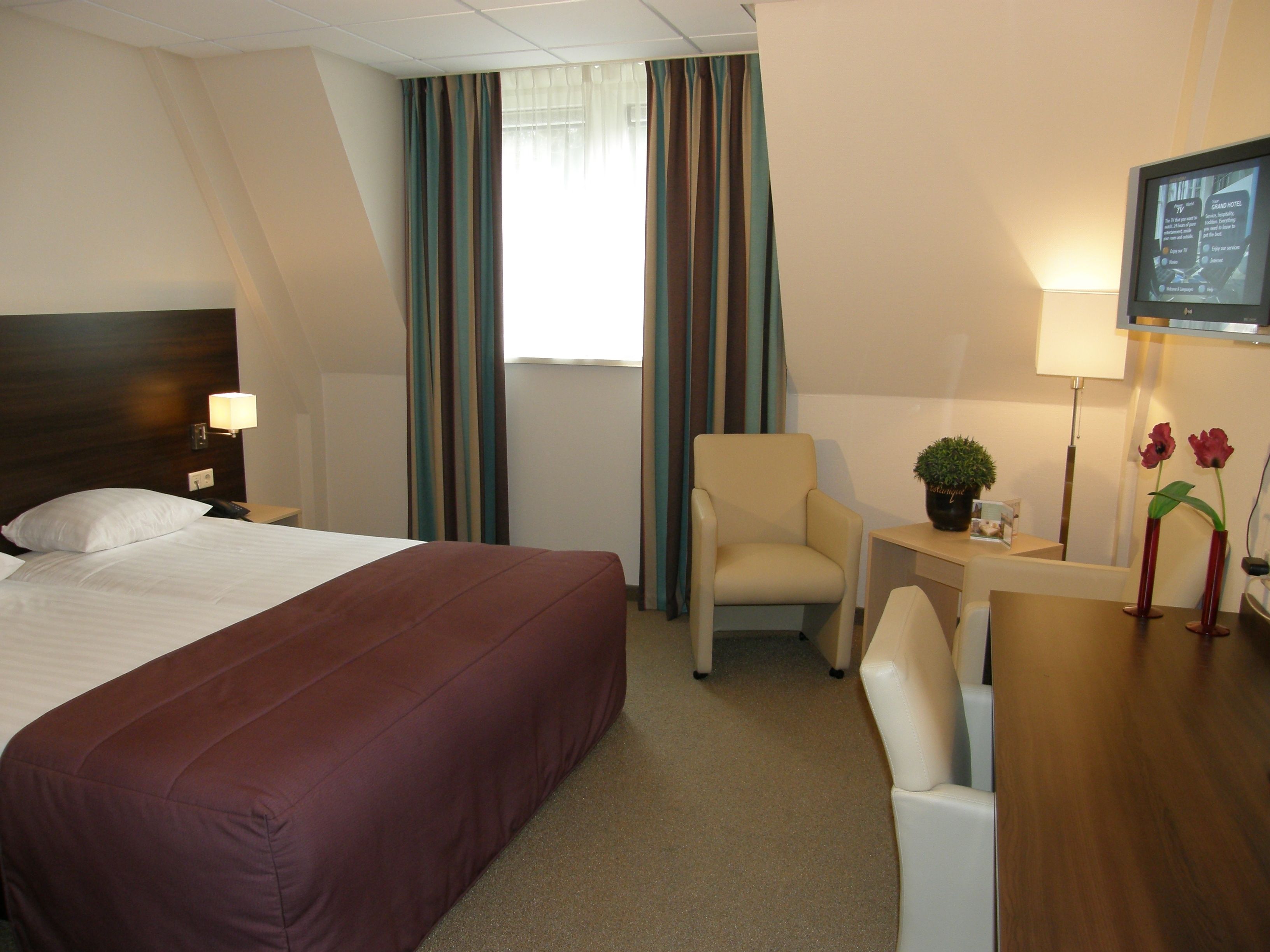 A hotel room with a large bed, a desk with a chair, a TV, and a window with curtains.