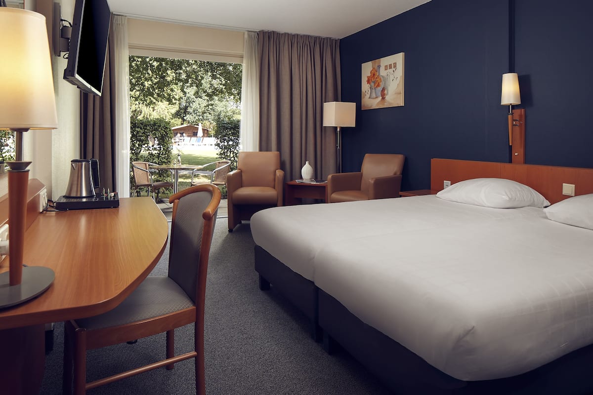 A hotel room with a bed, two armchairs, a desk with a lamp, a TV, and a painting on the wall.