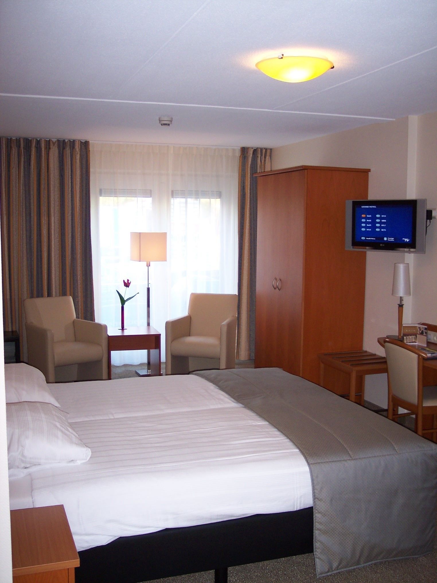 Una habitación de hotel con una cama grande, dos sillones, un escritorio, un televisor y una ventana con cortinas.