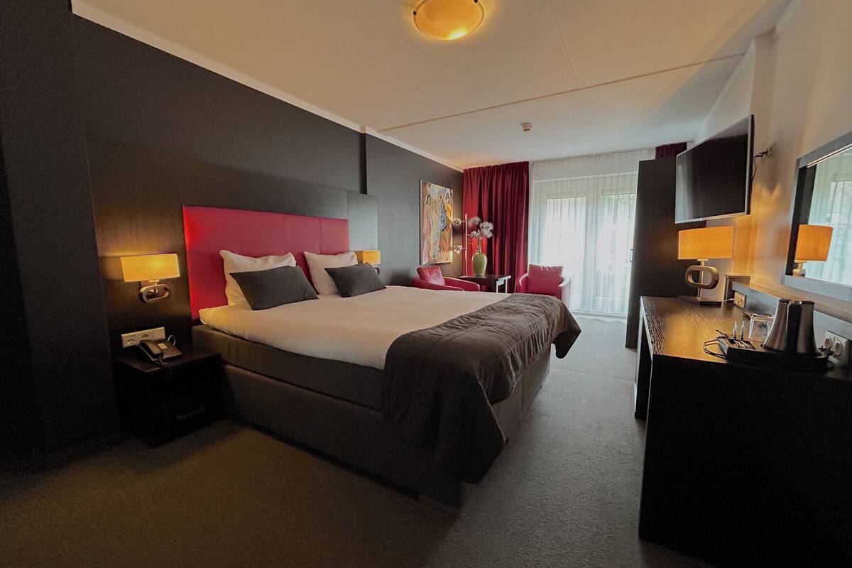 A hotel room with a large bed, bedside tables with lamps, a desk with a chair, a TV, and a window with curtains.