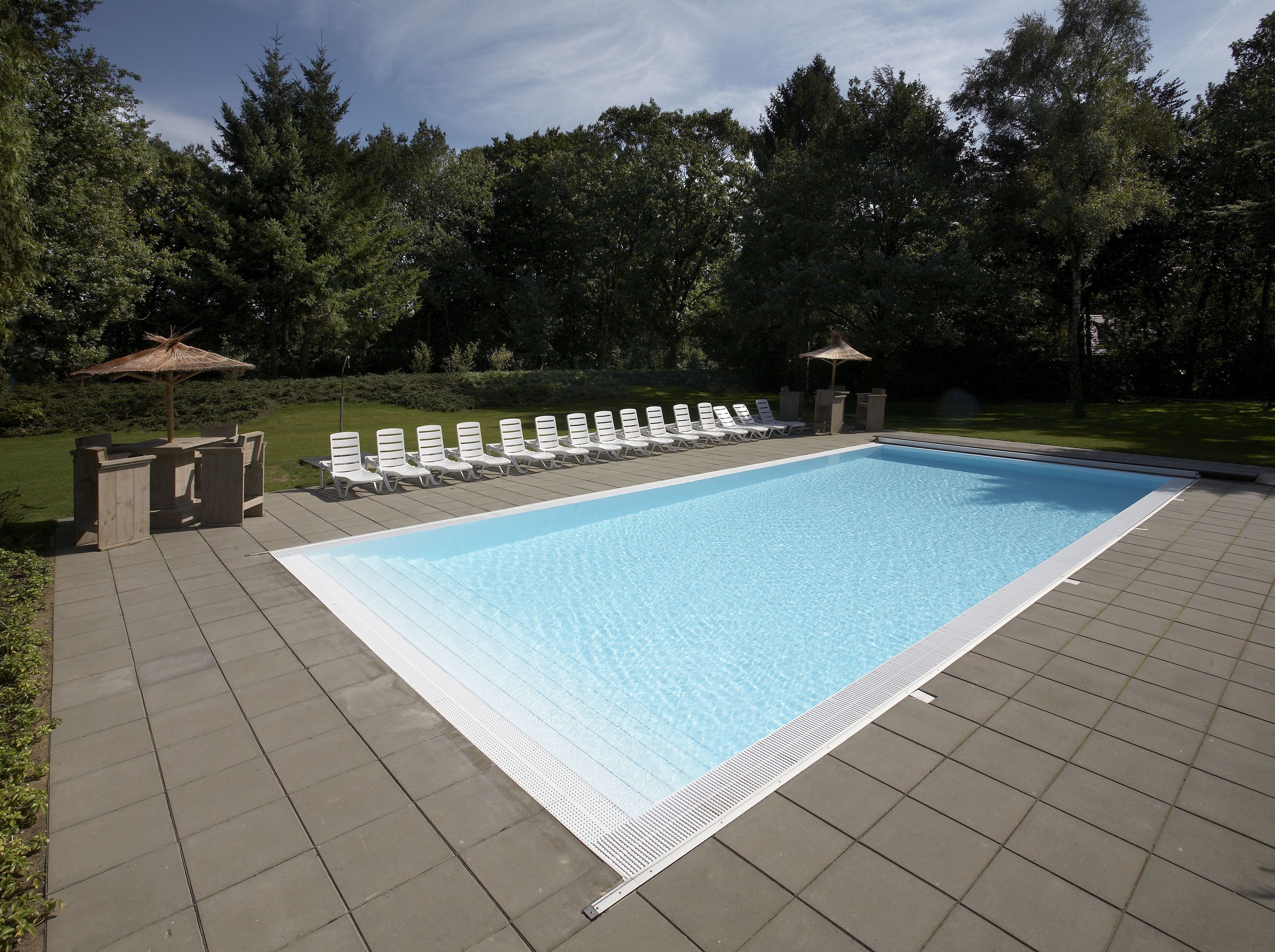 A rectangular swimming pool with a tiled deck, surrounded by white lounge chairs and two small umbrellas.