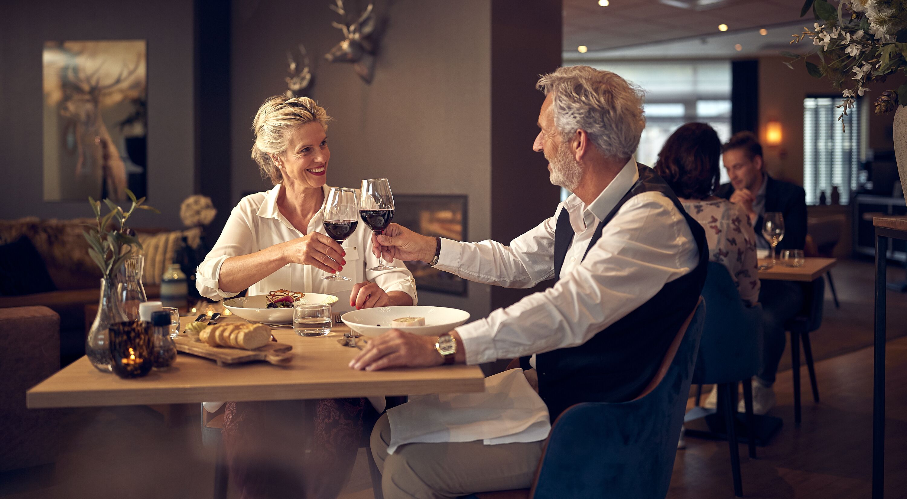 A couple toasting with wine glasses at a restaurant table with a candle, wine bottle, and bread.