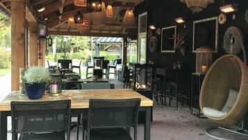 An outdoor dining area with wooden tables and chairs, hanging lanterns, and a wicker chair.