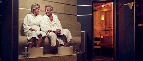 Dos personas con batas blancas sentadas en un banco en un entorno tipo spa, con interior de madera y pared de azulejos.