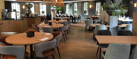 A modern restaurant with round tables and grey upholstered chairs, featuring a wooden floor and pendant lighting.