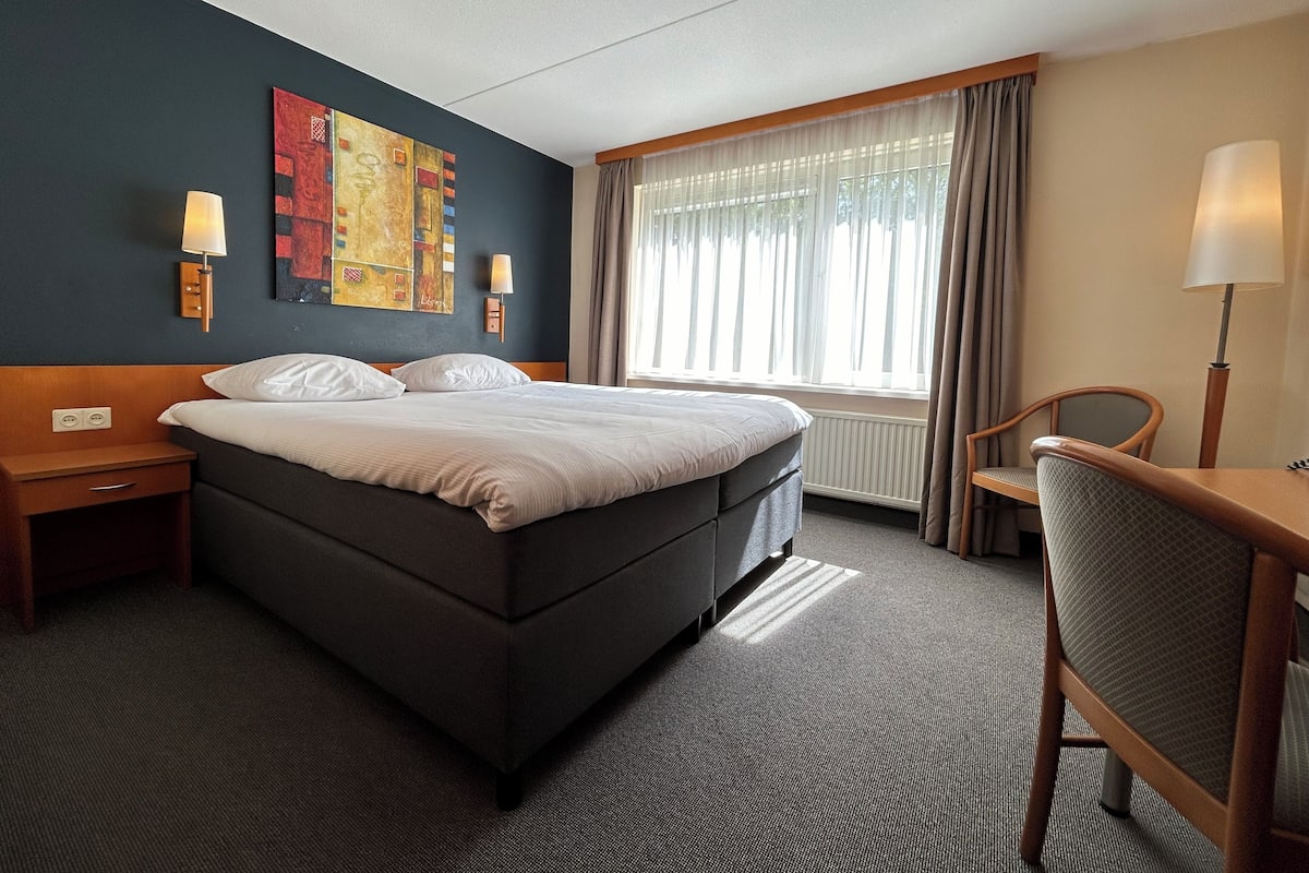 A hotel room with a large bed, two bedside tables, a desk with a chair, a window with curtains, and artwork on the wall.