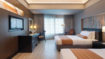 Habitación de hotel con dos camas, un escritorio con silla, televisión y un amplio ventanal con cortinas.