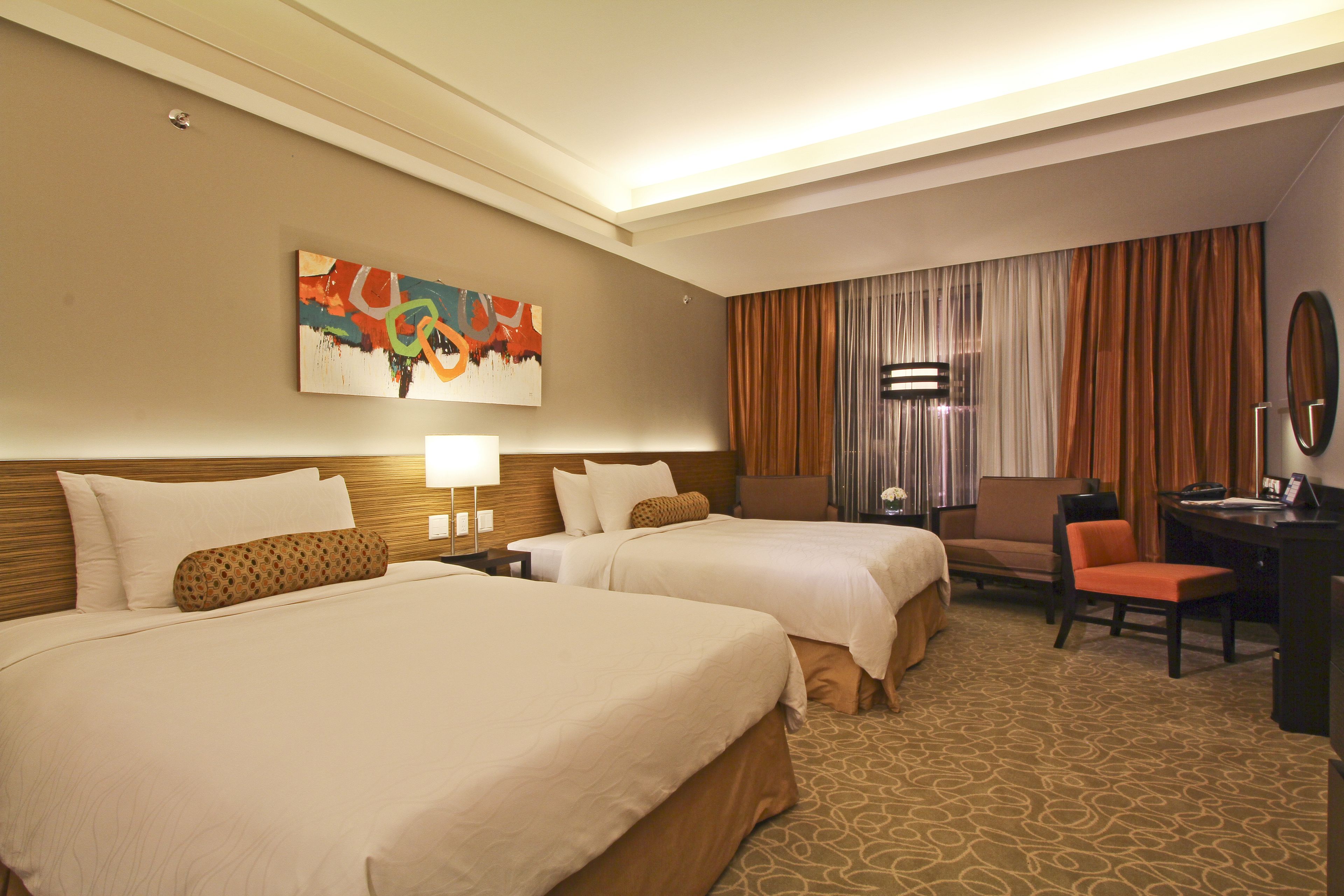 A hotel room with two beds, a desk, a chair, a lamp, and a painting on the wall.