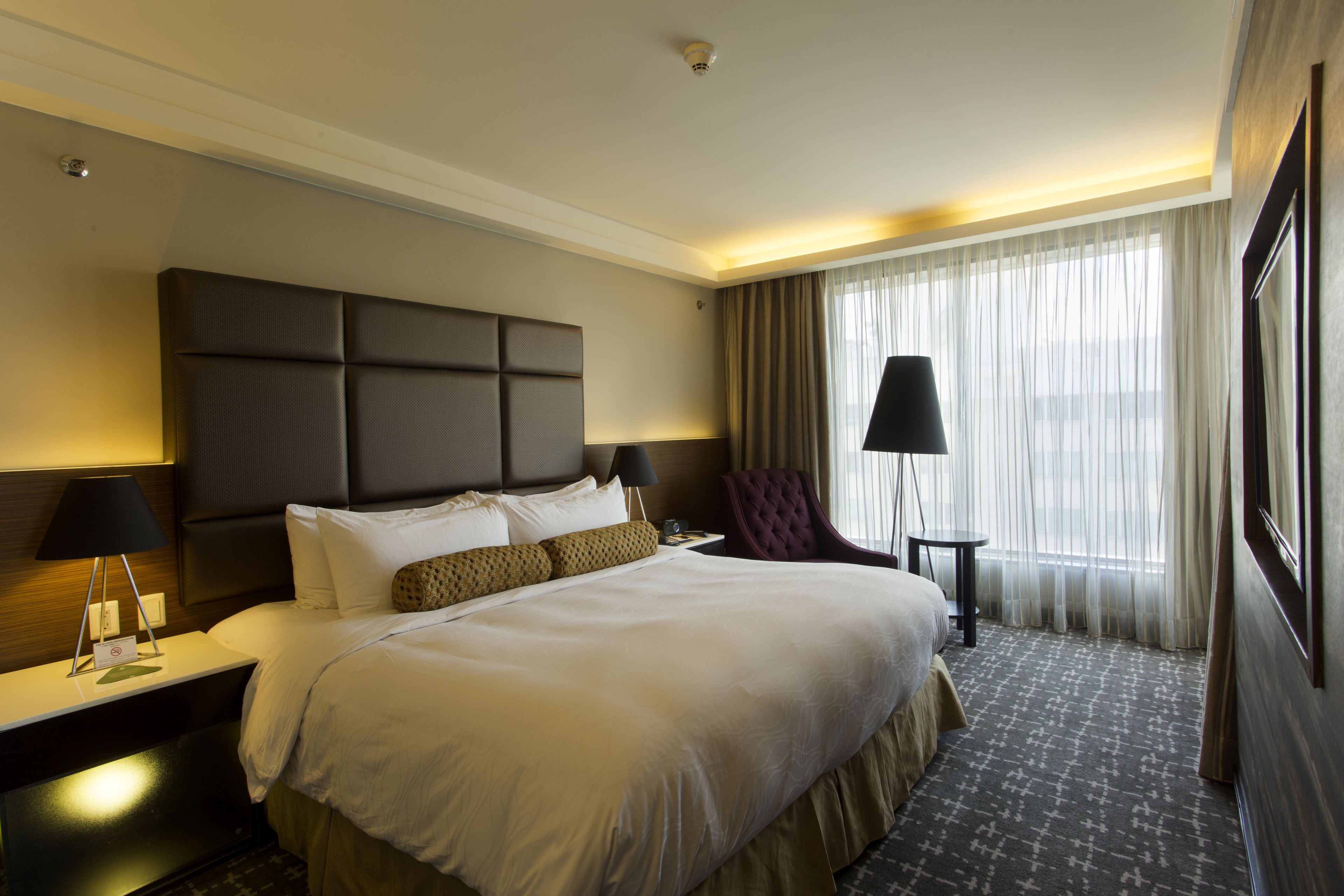 A hotel room with a large bed, a headboard, a nightstand, a chair, a lamp, and a window with curtains.