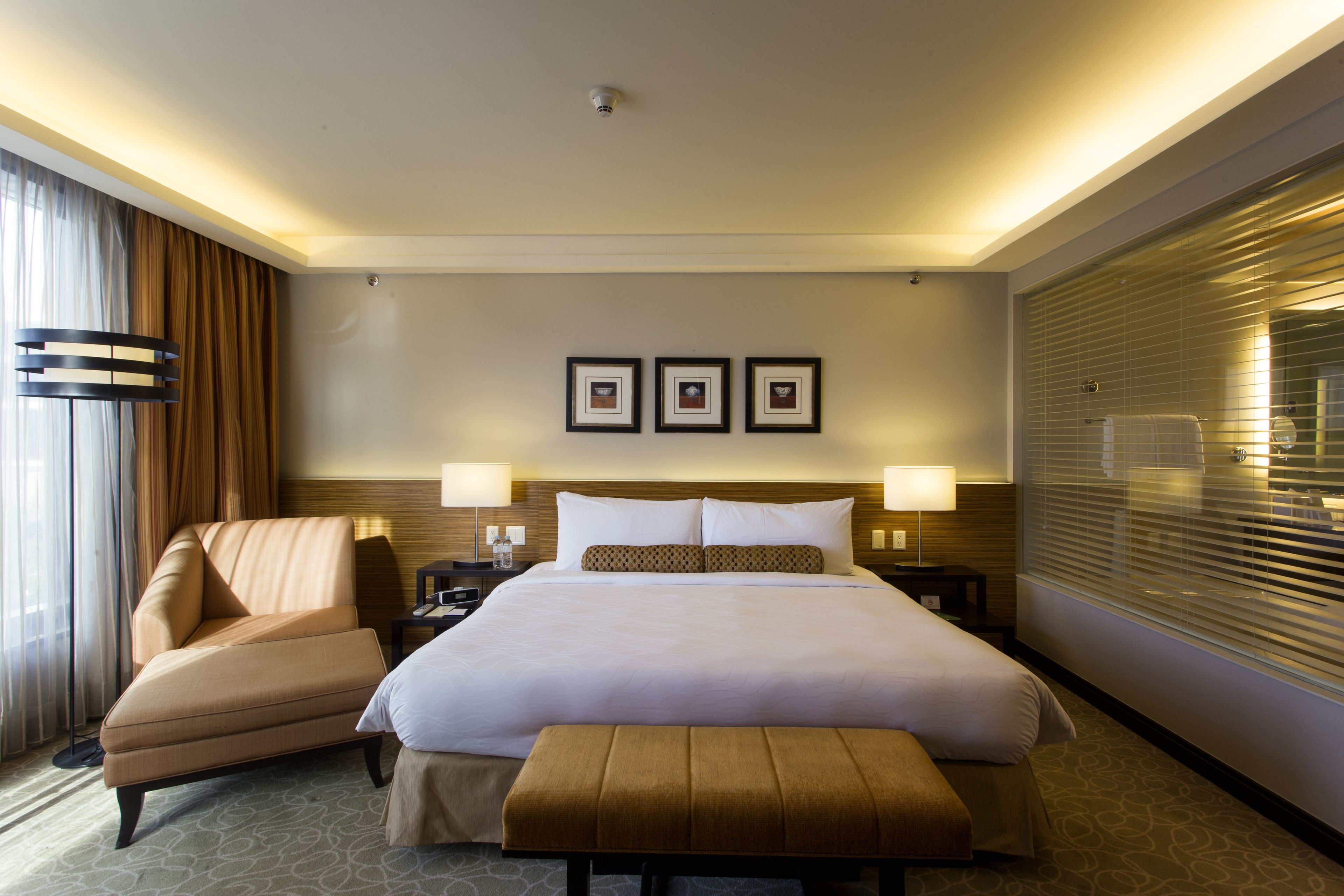 A hotel room with a large bed, a chair, a bench, and a nightstand with lamps.