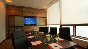 A conference room with a long table, chairs, a television screen, and a city view through large windows.