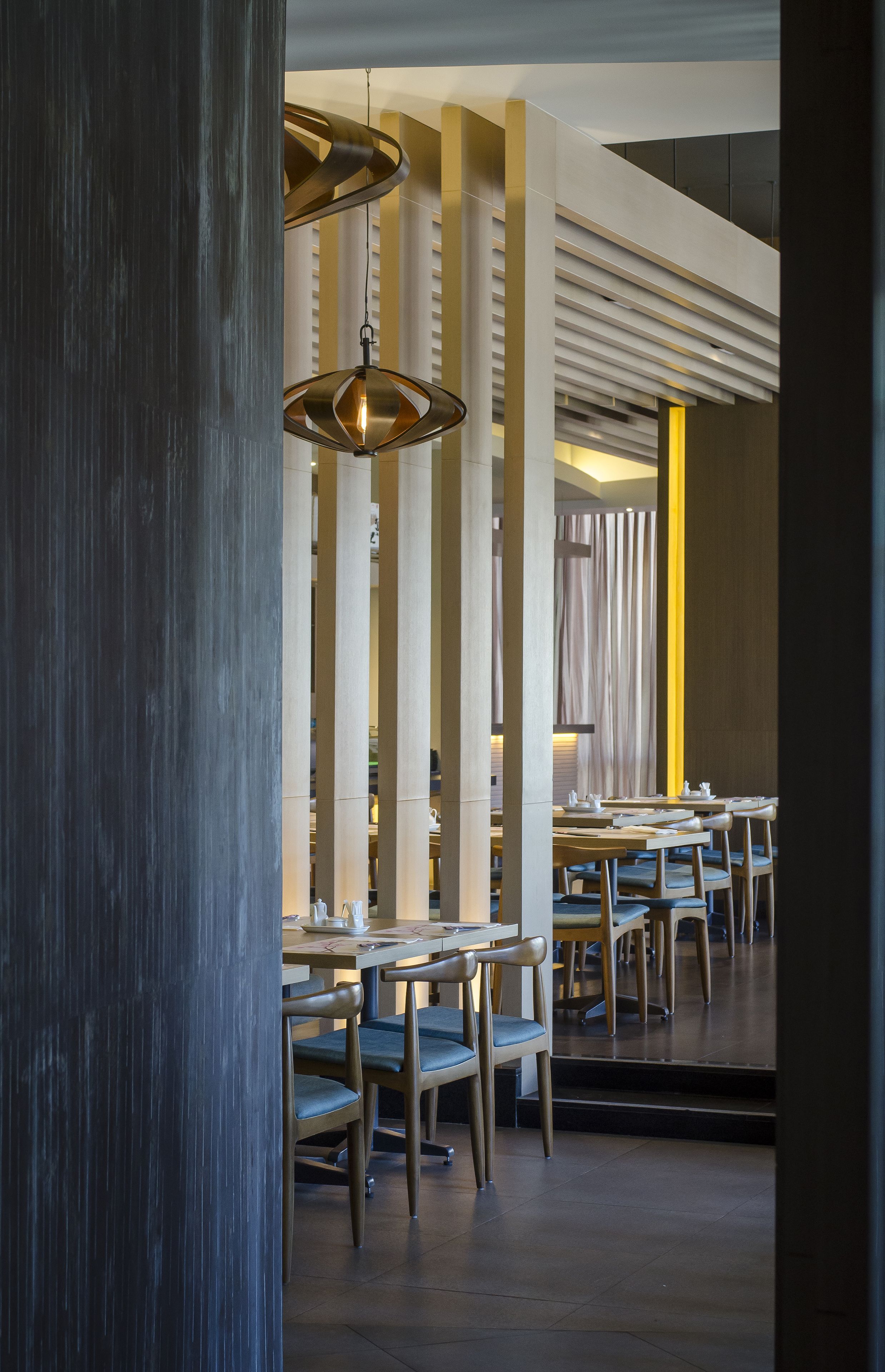 Interior de restaurante moderno con techos altos, columnas a la vista e iluminación colgante. Mesas dispuestas para comer.