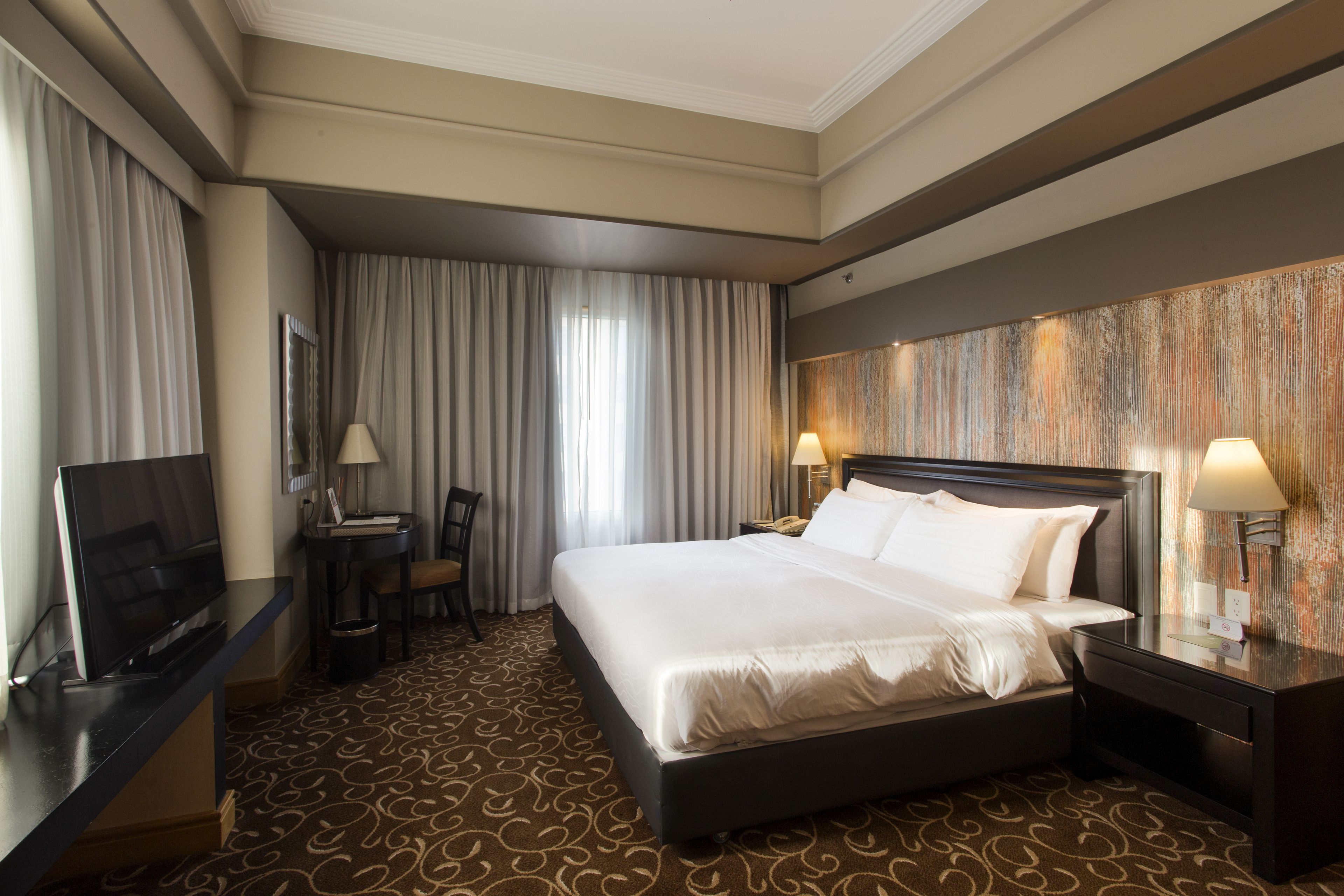 Una habitación de hotel con una cama grande, un escritorio con silla, un televisor y una lámpara fijada a la pared.