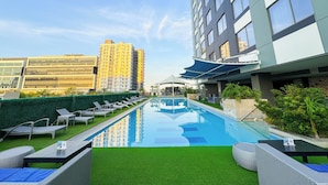 A rooftop pool with lounge chairs, a clear sky, and modern buildings in the background. - The Bellevue Manila  (Muntinlupa)