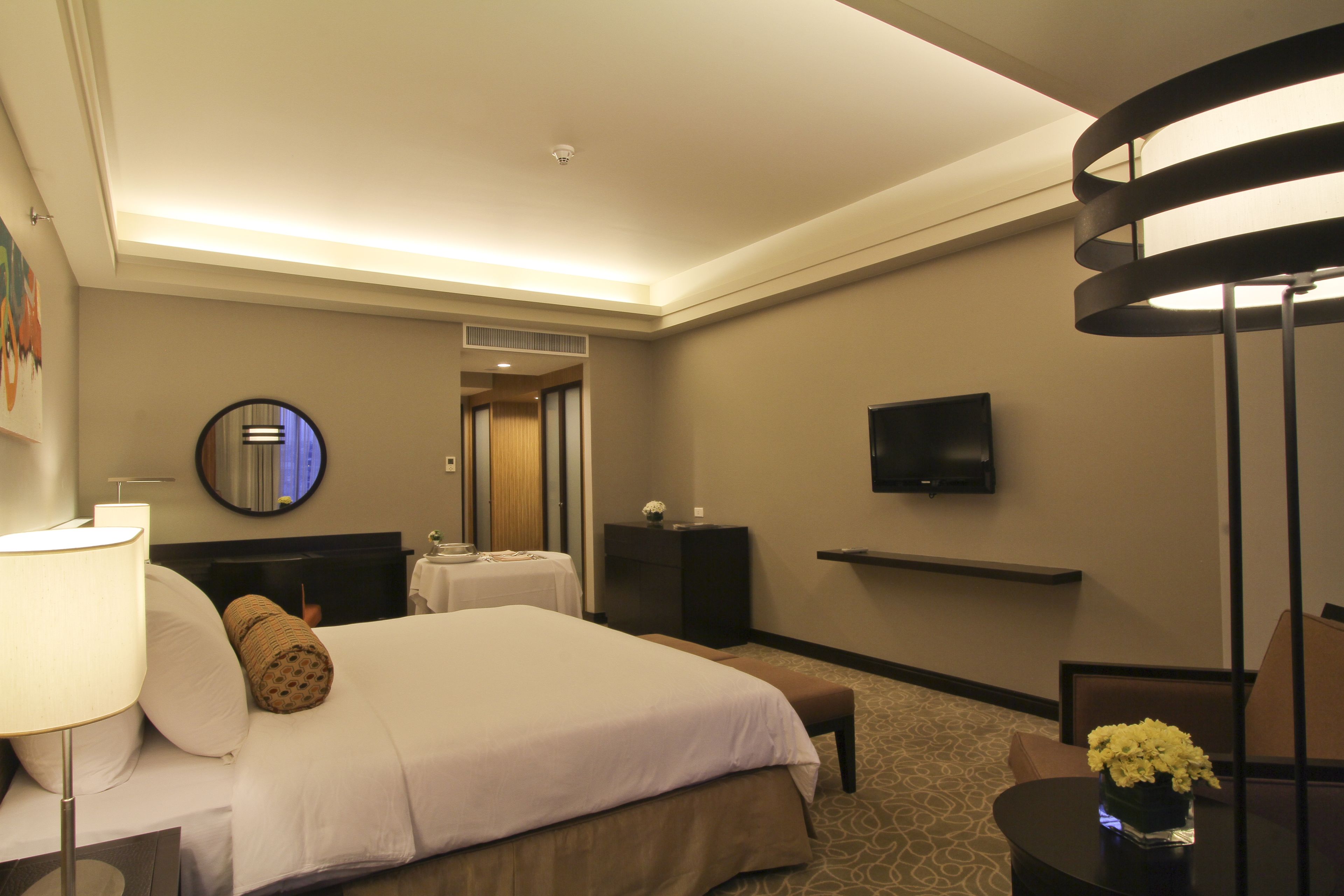 Una habitación de hotel con una cama grande, mesitas de noche, un televisor de pantalla plana, un espejo redondo y una pequeña mesa con un florero.