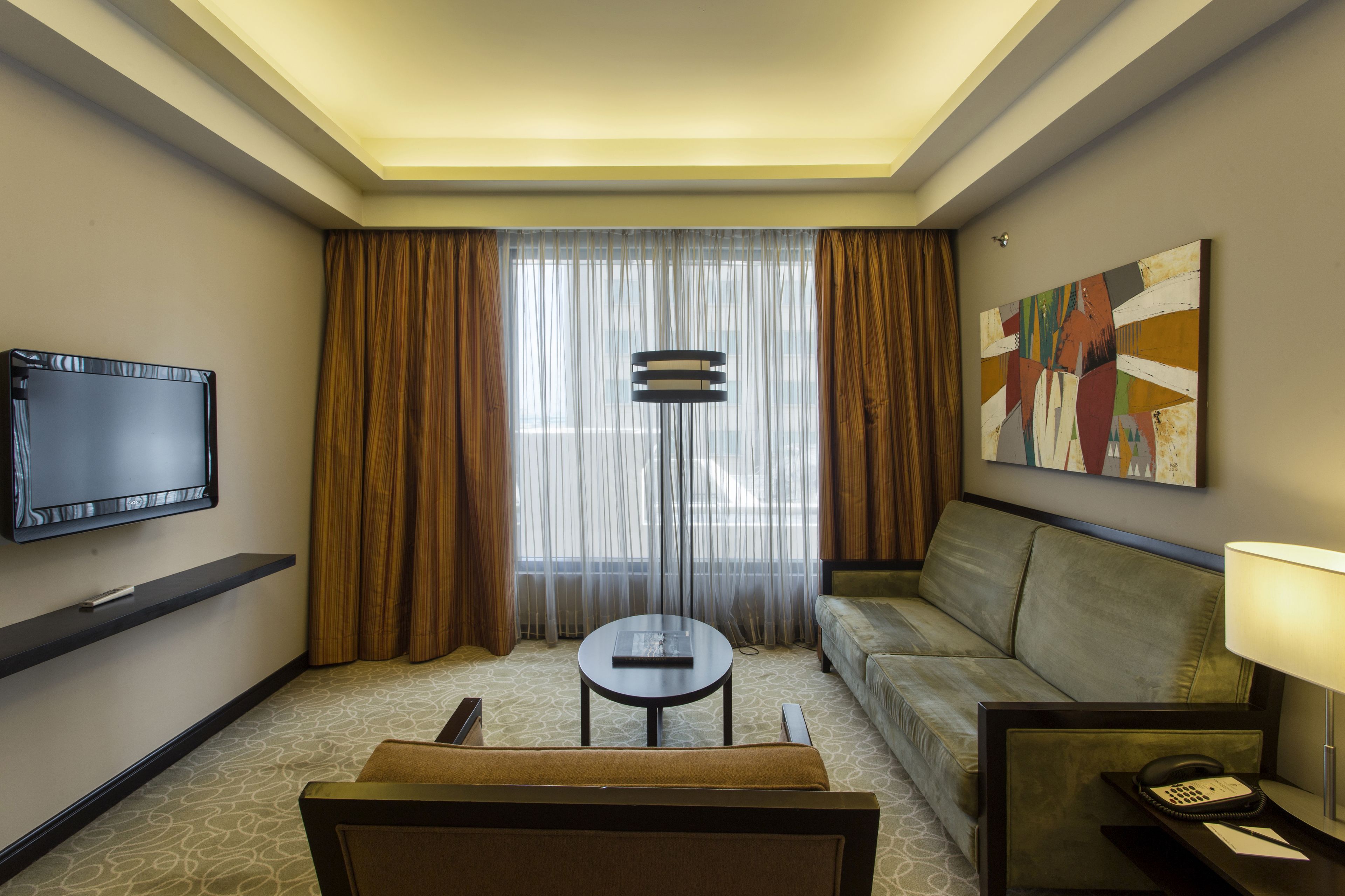 A hotel room with a flat-screen TV, a sofa, a small round table, a lamp, and a painting on the wall.