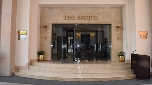 A entrada do hotel The Bristol, com fachada de mármore, degraus que levam à entrada e duas plantas em vasos de cada lado.