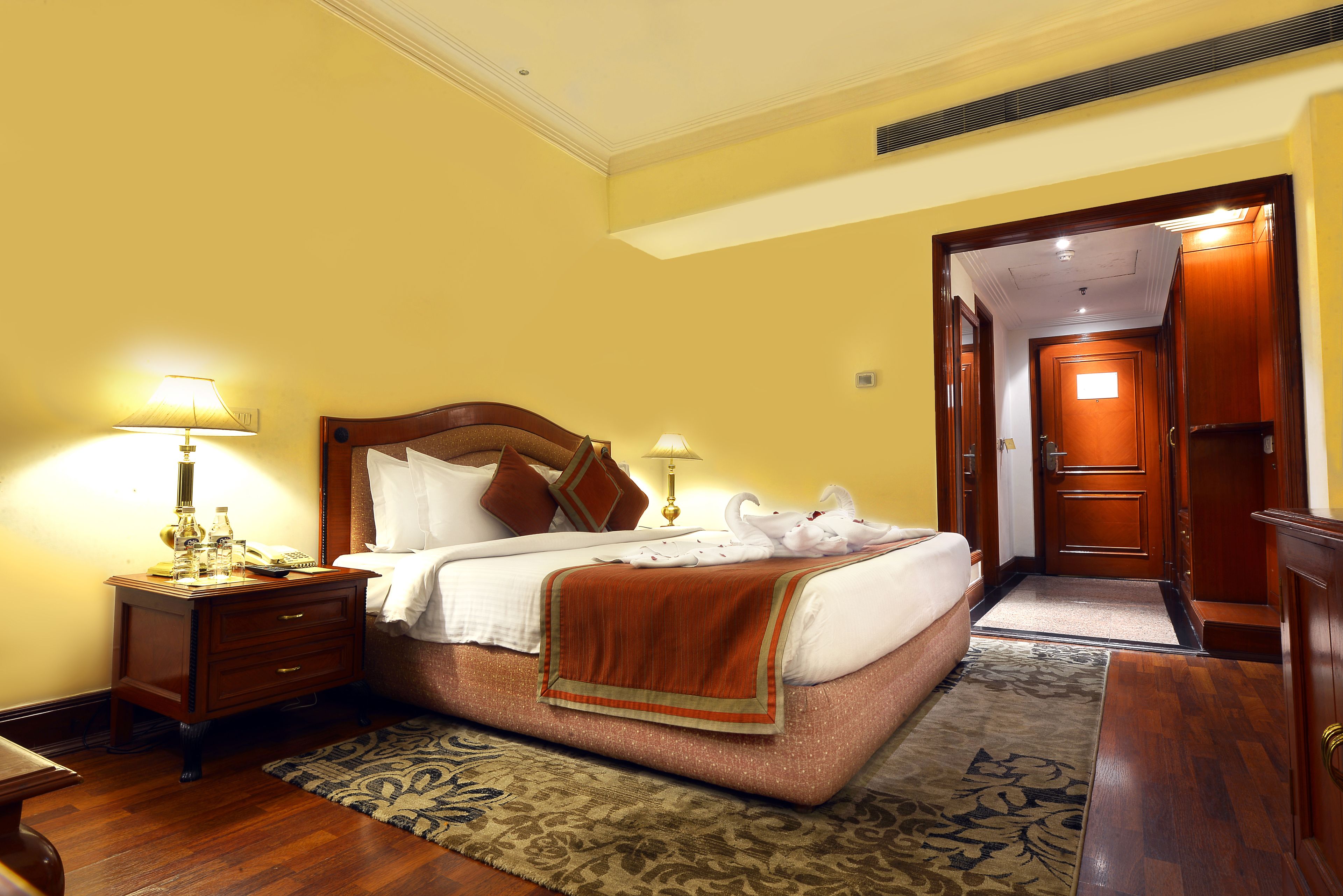 A hotel room with a bed, wooden nightstand, lamps, and a door leading to another room.