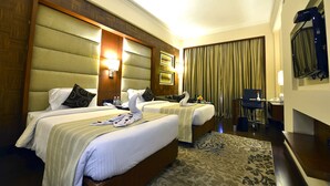 Quarto de hotel com duas camas, uma escrivaninha, uma cadeira, televisão e janela com cortinas.