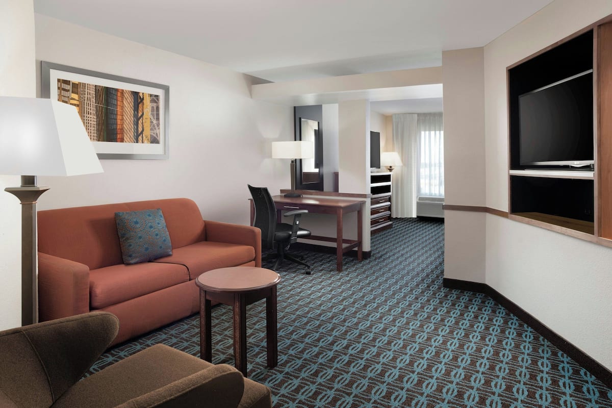 A hotel room with a sofa, a small table, a desk, a chair, a TV, and a patterned carpet.