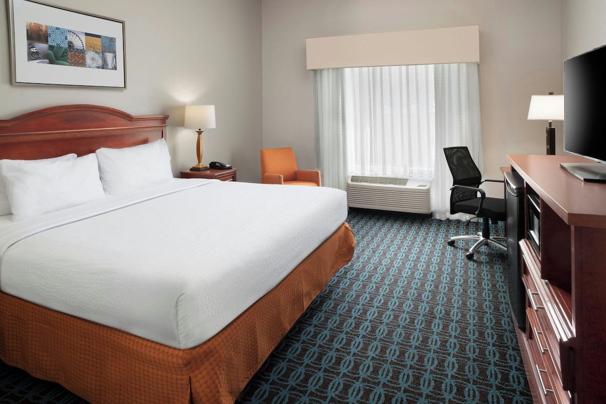 A hotel room with a large bed, a desk with a television, an air conditioning unit, and a chair.